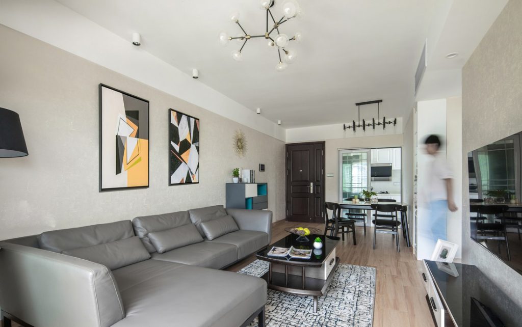 تحتوي غرفة المعيشة على أريكة كبيرة على شكل حرف L وطاولة شاي خشبية صغيرة مع زجاج أسود ولوحات زخرفية ورف تلفزيون